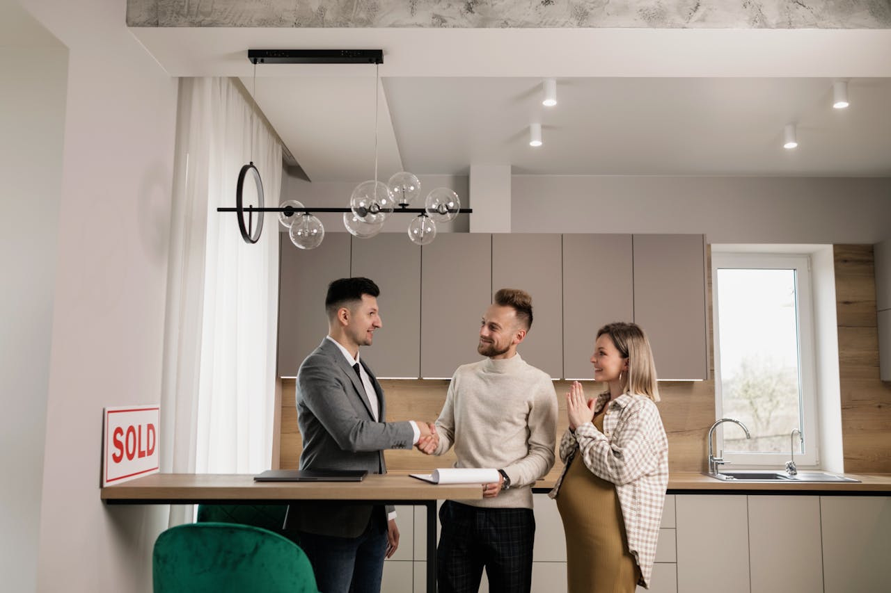 A smiling couple and a realtor finalize a house purchase in a modern kitchen setting, sealing the deal with a handshake.